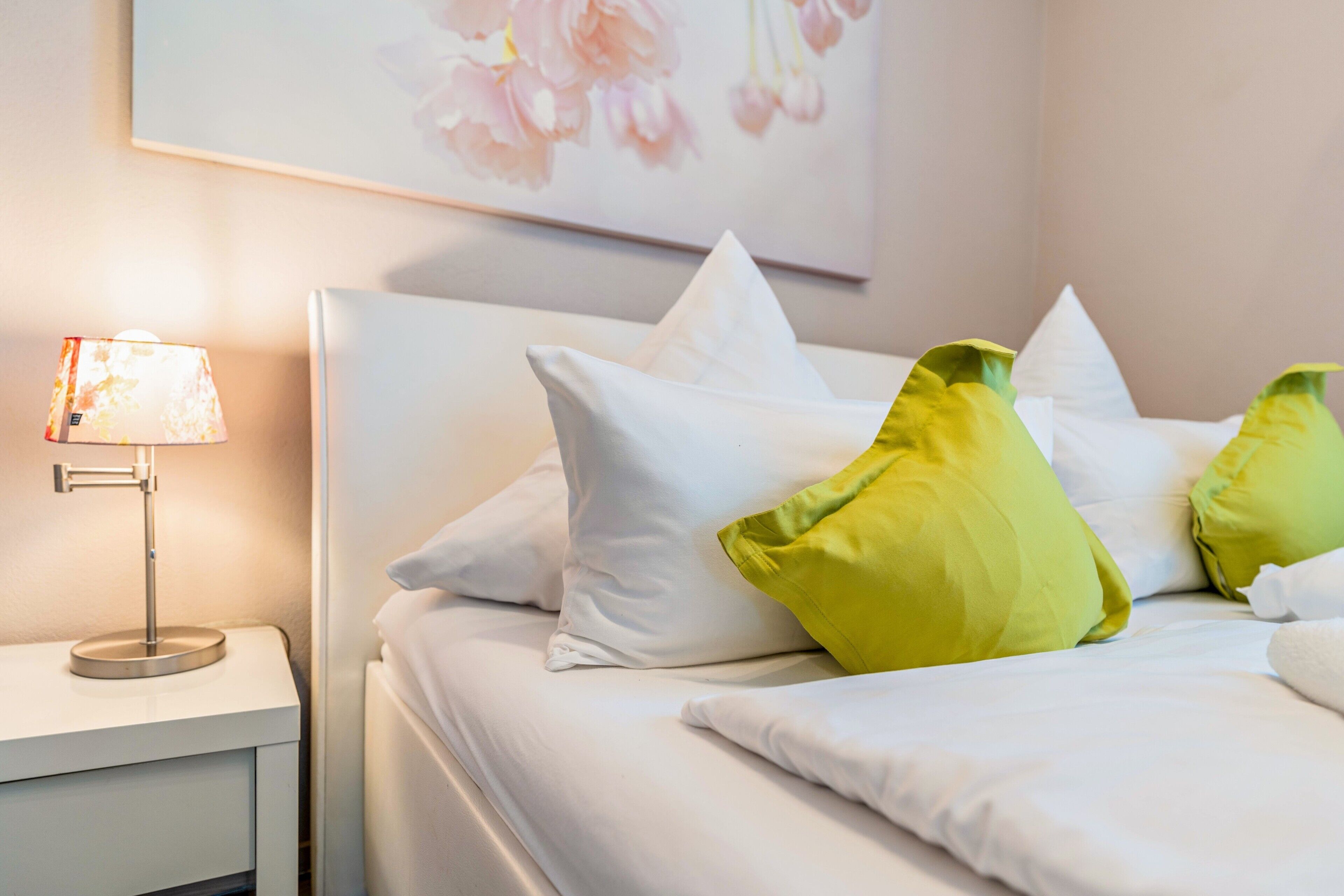 Ein ordentlich bezogenes Bett mit weißen und grünen Kissen, einer Nachttischlampe und einer Wand mit einem Blumenbild.