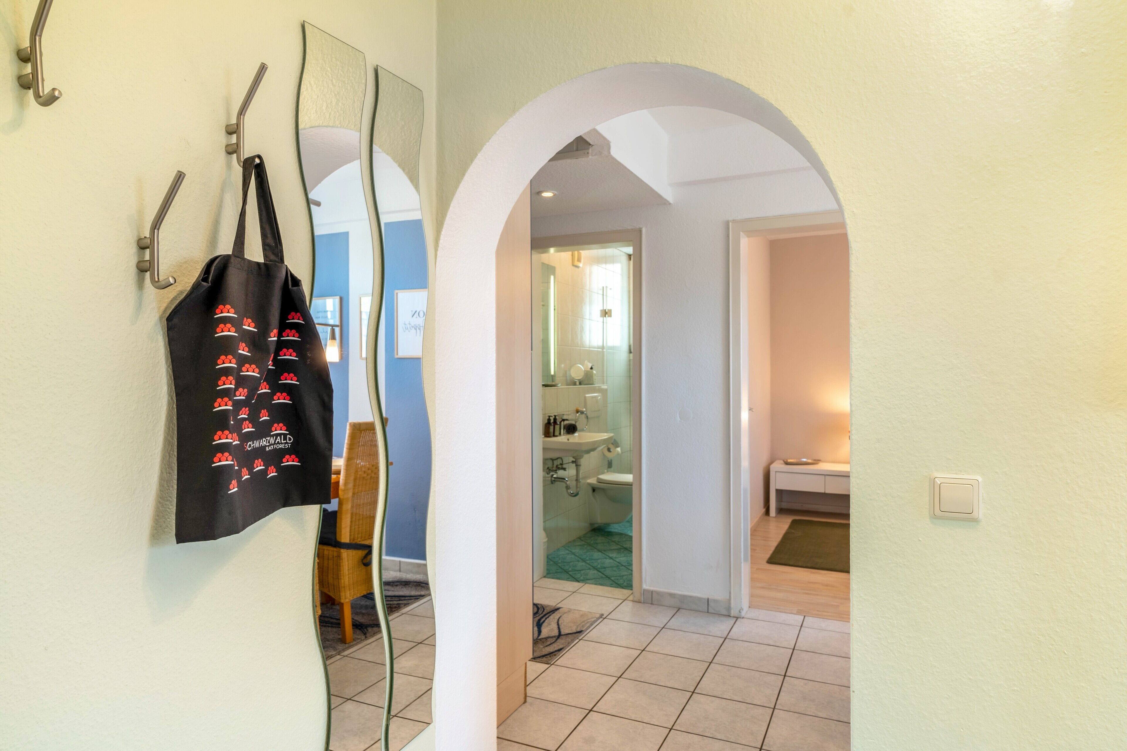 A hallway with a mirror, a black tote bag with red cherries, and a bathroom visible through an archway.