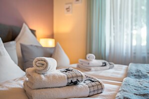 Una cama bien hecha con toallas enrolladas y una manta escocesa, una lámpara de noche y una ventana con cortinas.