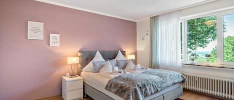 A bedroom with a bed, bedside tables, a window with curtains, and a view of greenery outside.