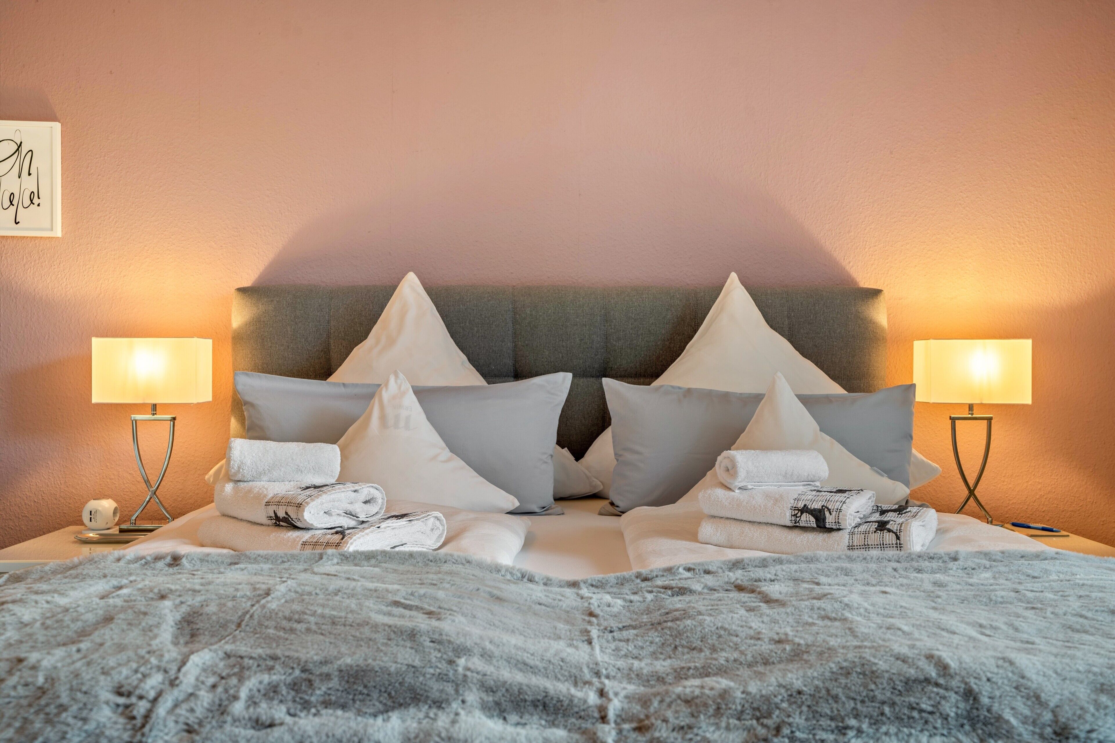 A neatly made bed with multiple pillows and folded towels, two bedside lamps, and a wall-mounted artwork.