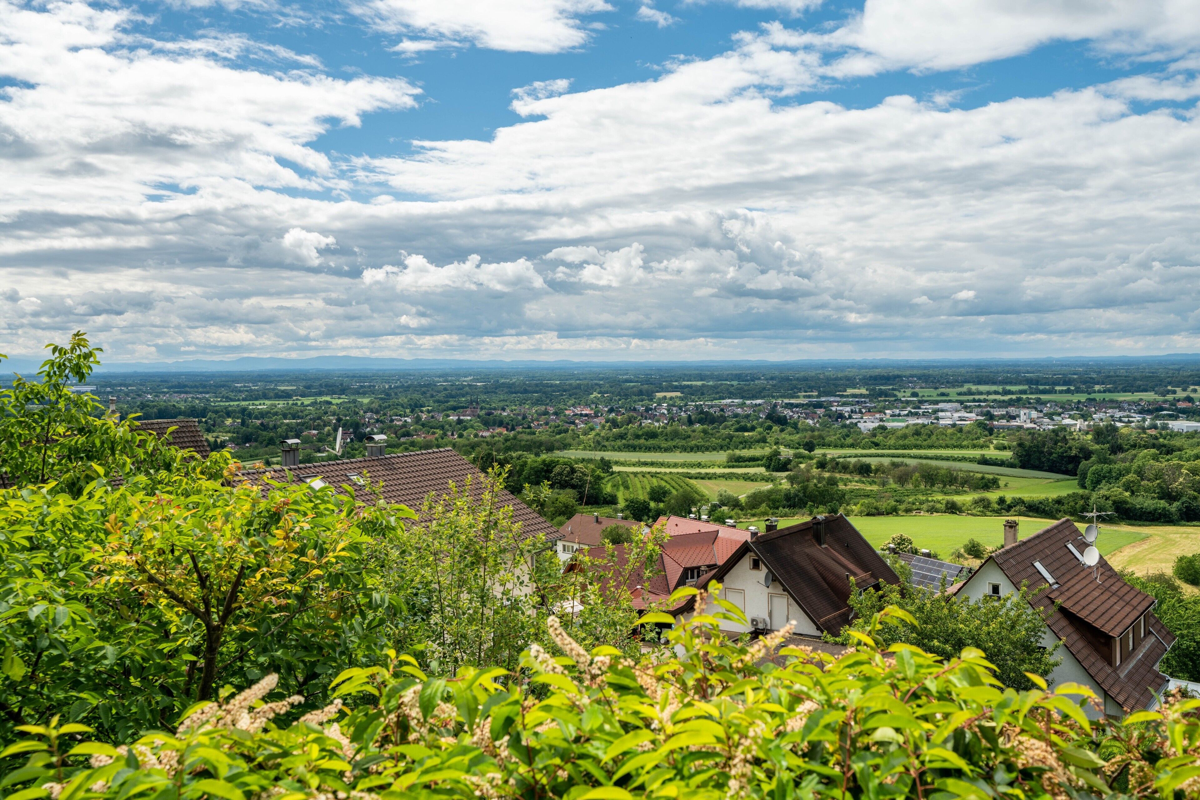 Ländliche Landschaft mit Häusern, viel Grün und einem bewölkten Himmel.