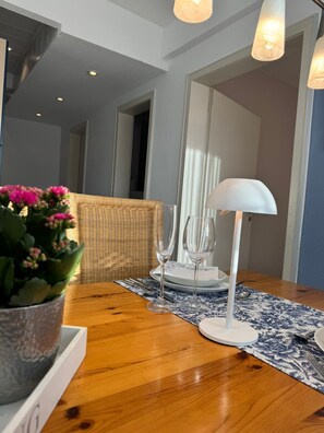 Ein Esstisch mit einer Topfpflanze, zwei Weingläsern, einer weißen Tischlampe und einem Korbstuhl.