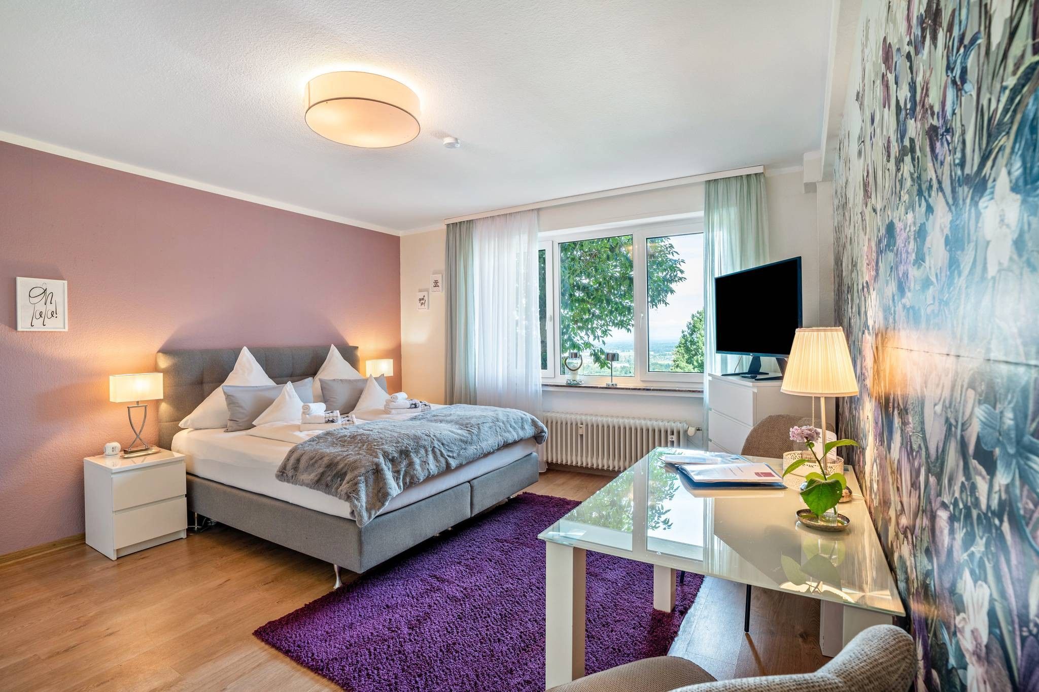 A modern hotel room with a bed, bedside tables, a desk, a TV, and a window with a view.