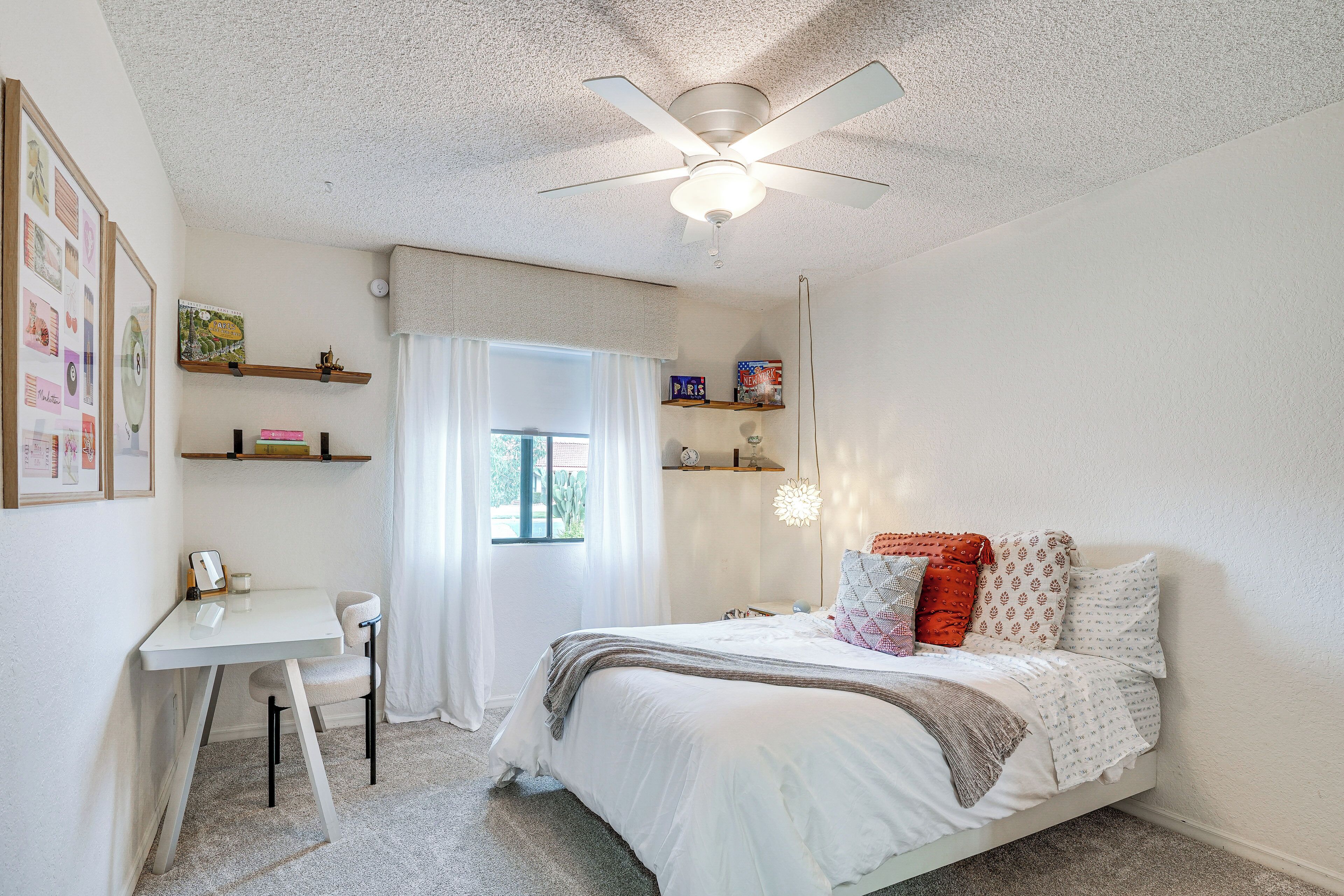 A bedroom with a bed, a desk, and a chair. There is a ceiling fan, a window with curtains, and shelves with items on them.