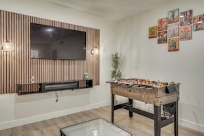 A living room with a foosball table, a flat-screen TV mounted on a wooden panel, and a wall with framed artwork. - Summer-ready Old Town Condo w/ Pool & Patio Access (Scottsdale)