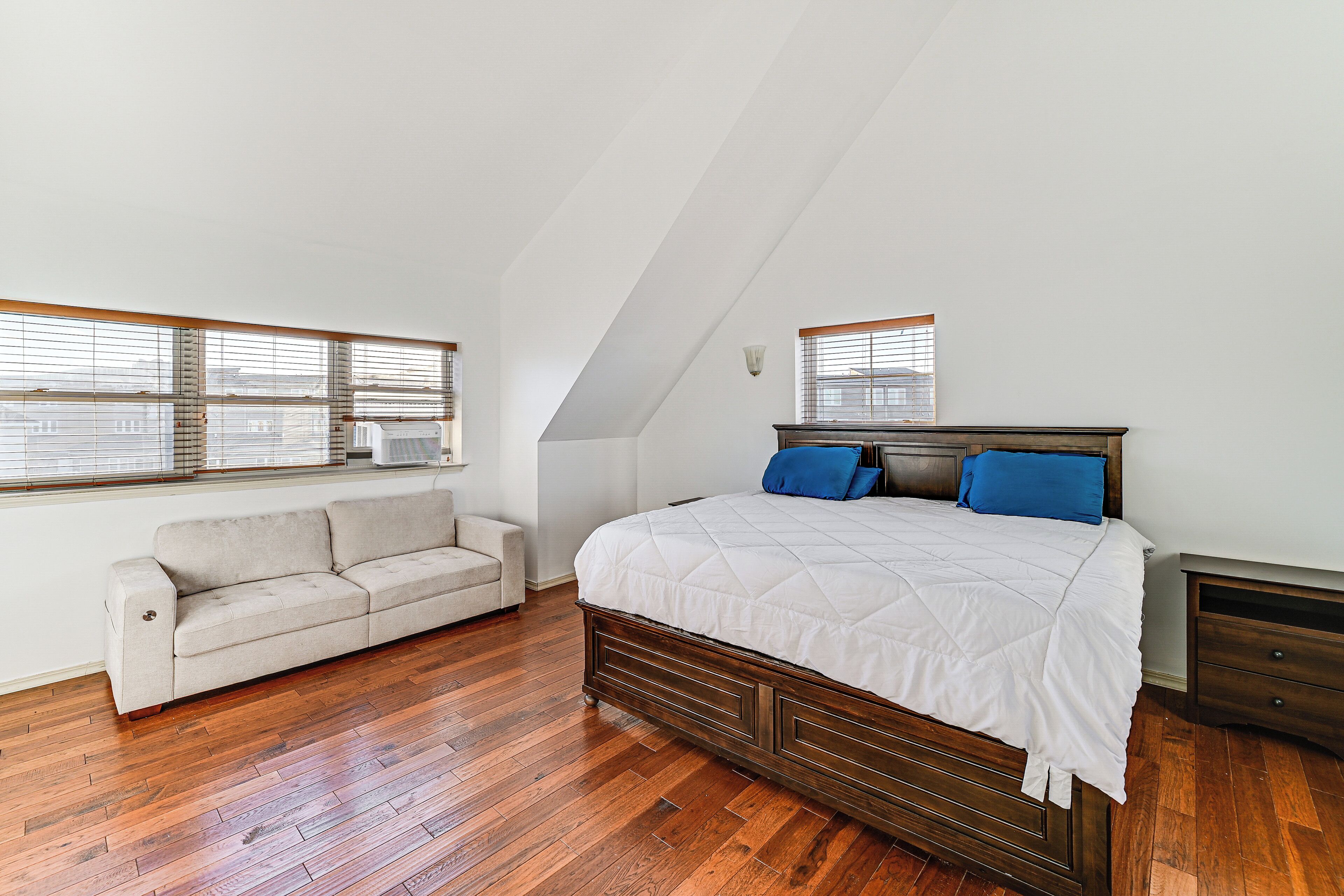 A bedroom with a wooden floor, a bed with a white duvet, a beige sofa, and two windows with blinds.