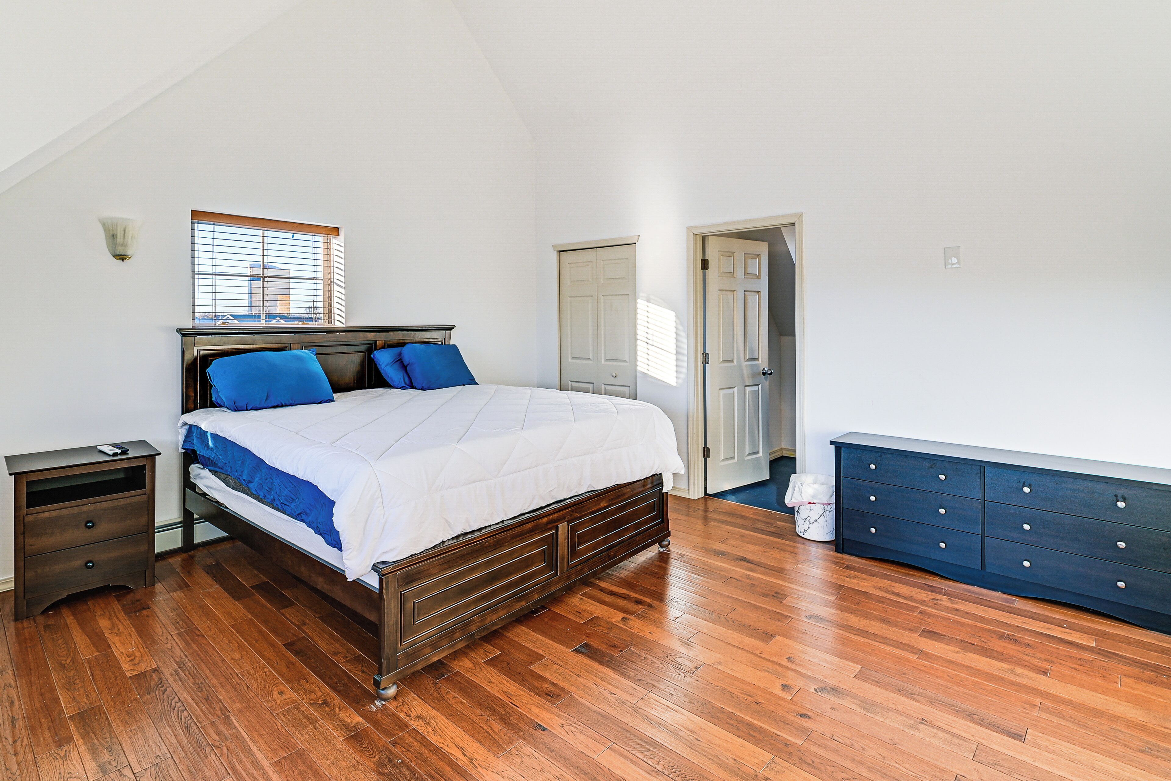 A bedroom with a wooden floor, a bed with blue bedding, a nightstand, a dresser, and a window with blinds.