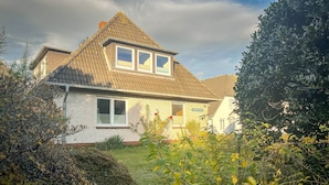 A two-story house with a gabled roof, a visible chimney, and a front garden with various plants and shrubs. - Vacation apartment Eldorado Laacht (Norddorf)