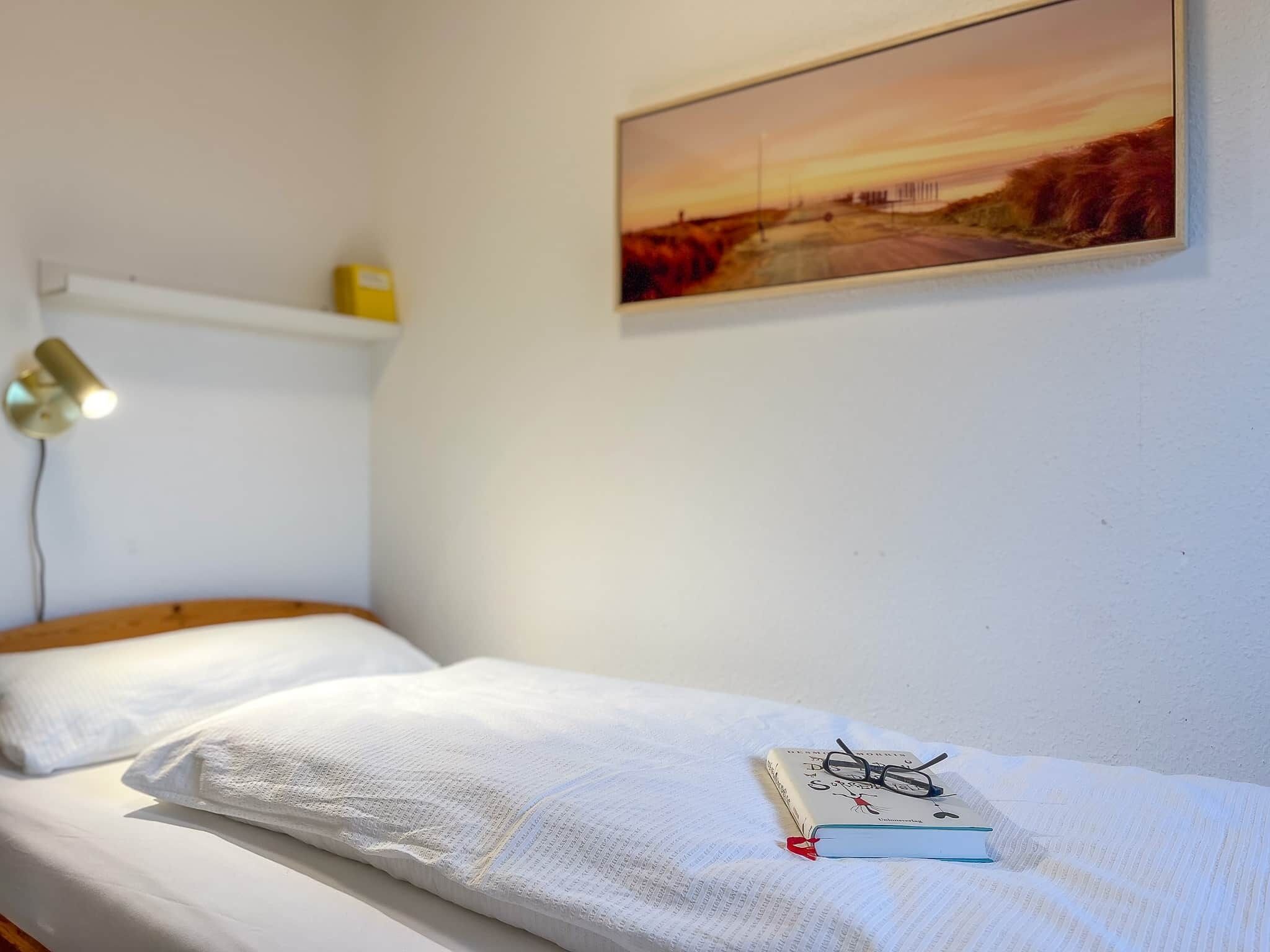A single bed with a white mattress and pillows, a bedside table with a lamp, a book, and glasses on the bed, and a painting of a sunset on the wall.