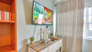 Una habitación con una estantería de madera, una consola blanca con adornos, un cuadro de una escena junto al lago y una ventana con cortinas.