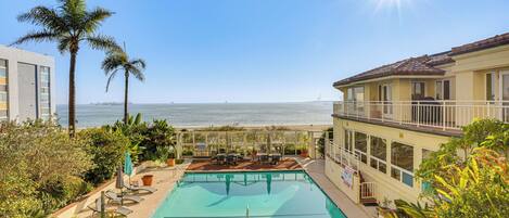 A pool with a view of the ocean, surrounded by palm trees and a building with a balcony.
