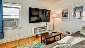 Een woonkamer met een flatscreen televisie, een witte televisiemeubel, een salontafel, een bank, een lamp en een stadsgezicht poster.