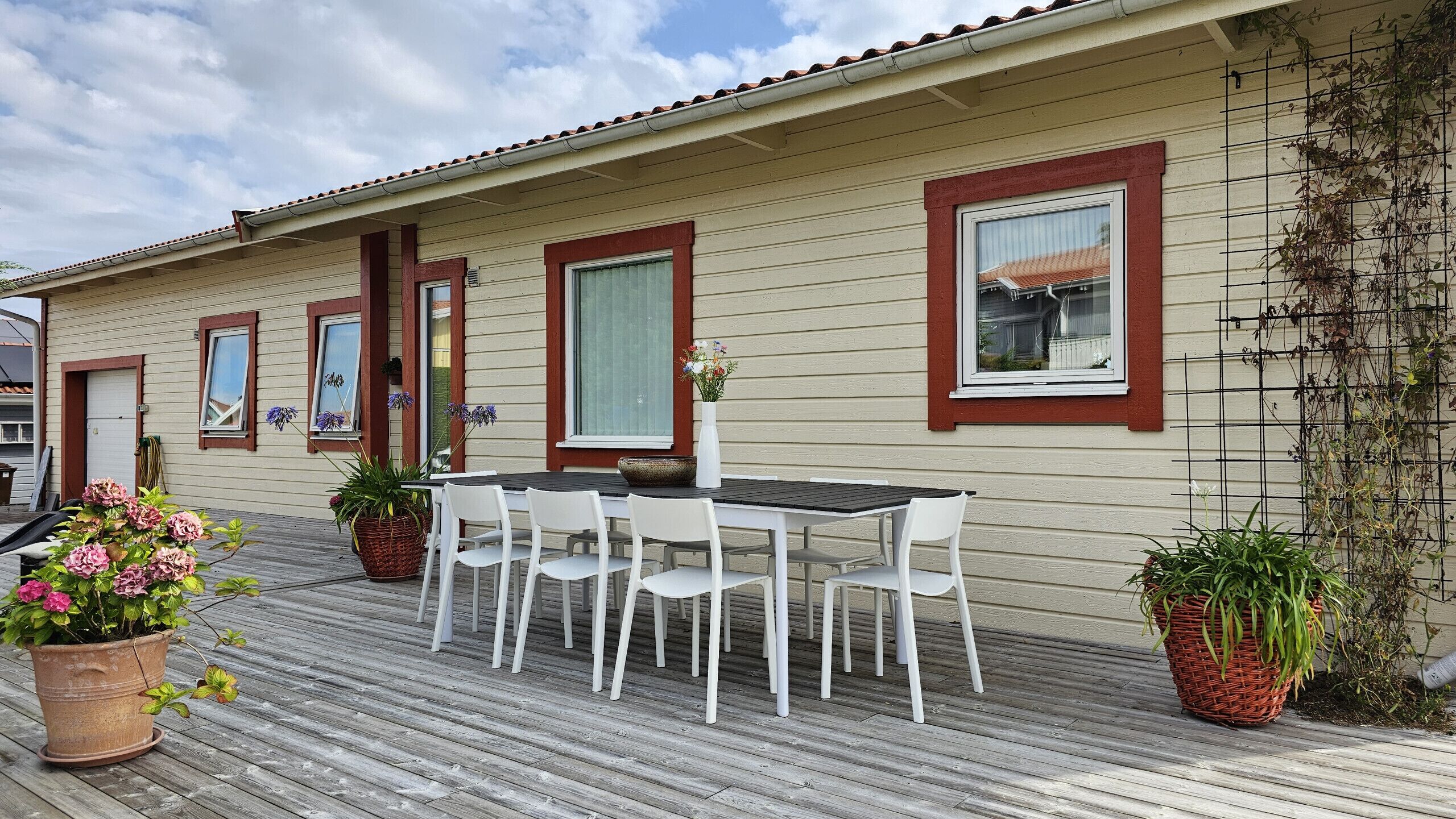 Eine Holzterrasse mit Tisch und Stühlen, Topfpflanzen und einem Haus mit großen Fenstern und roter Fassade.