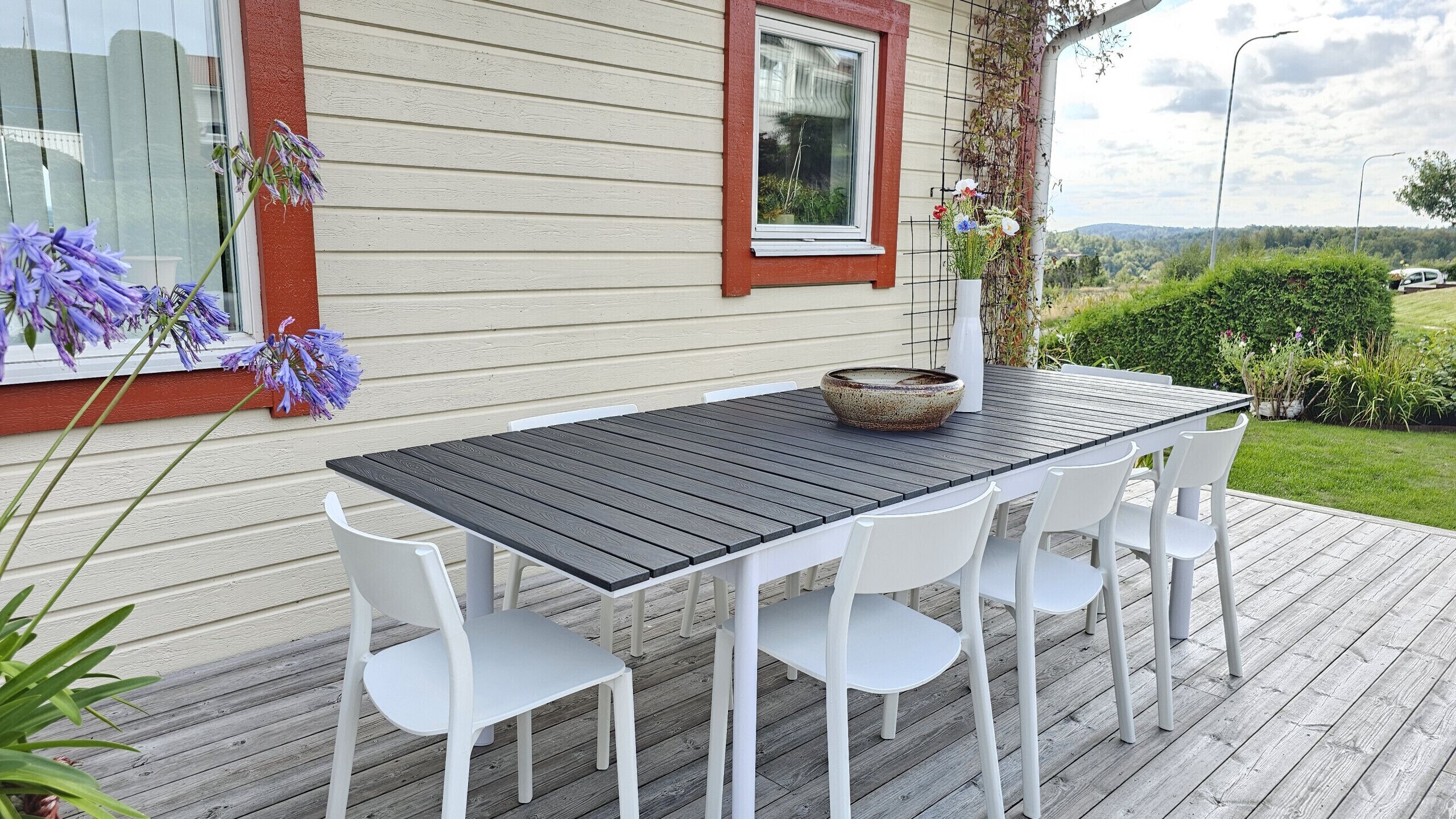 A wooden deck with a table and chairs, a window with a red frame, and a view of greenery and hills in the background.