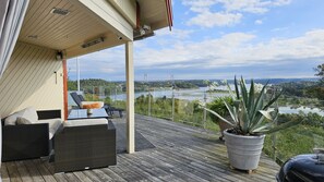Een overdekt terras met uitzicht op een rivier en een brug, ingericht met rieten meubels en een potplant.