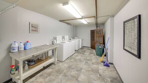 A laundry room with a washing machine, dryer, ironing board, and shelves with cleaning supplies. - Walk to Chair Lift: Mtn-view Schweitzer Studio! (Sandpoint)