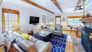 A living room with a sofa, coffee table, and fireplace. There is a television mounted on the wall and a window with blinds. - 4BR House, Steps to Beach w/ Deck, W & D & Puzzles (North Truro)
