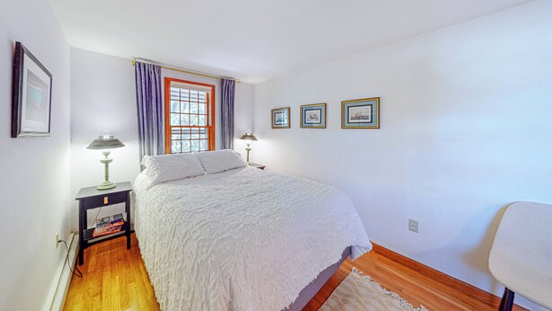 A bedroom with a bed, a nightstand with a lamp, a window with curtains, and framed pictures on the wall. - 4BR House, Steps to Beach w/ Deck, W & D & Puzzles (North Truro)