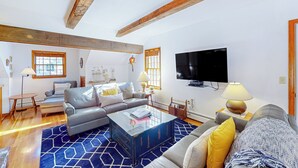 A spacious living room with a modern sofa set, a coffee table, and a flat-screen TV mounted on the wall. - 4BR House, Steps to Beach w/ Deck, W & D & Puzzles (North Truro)