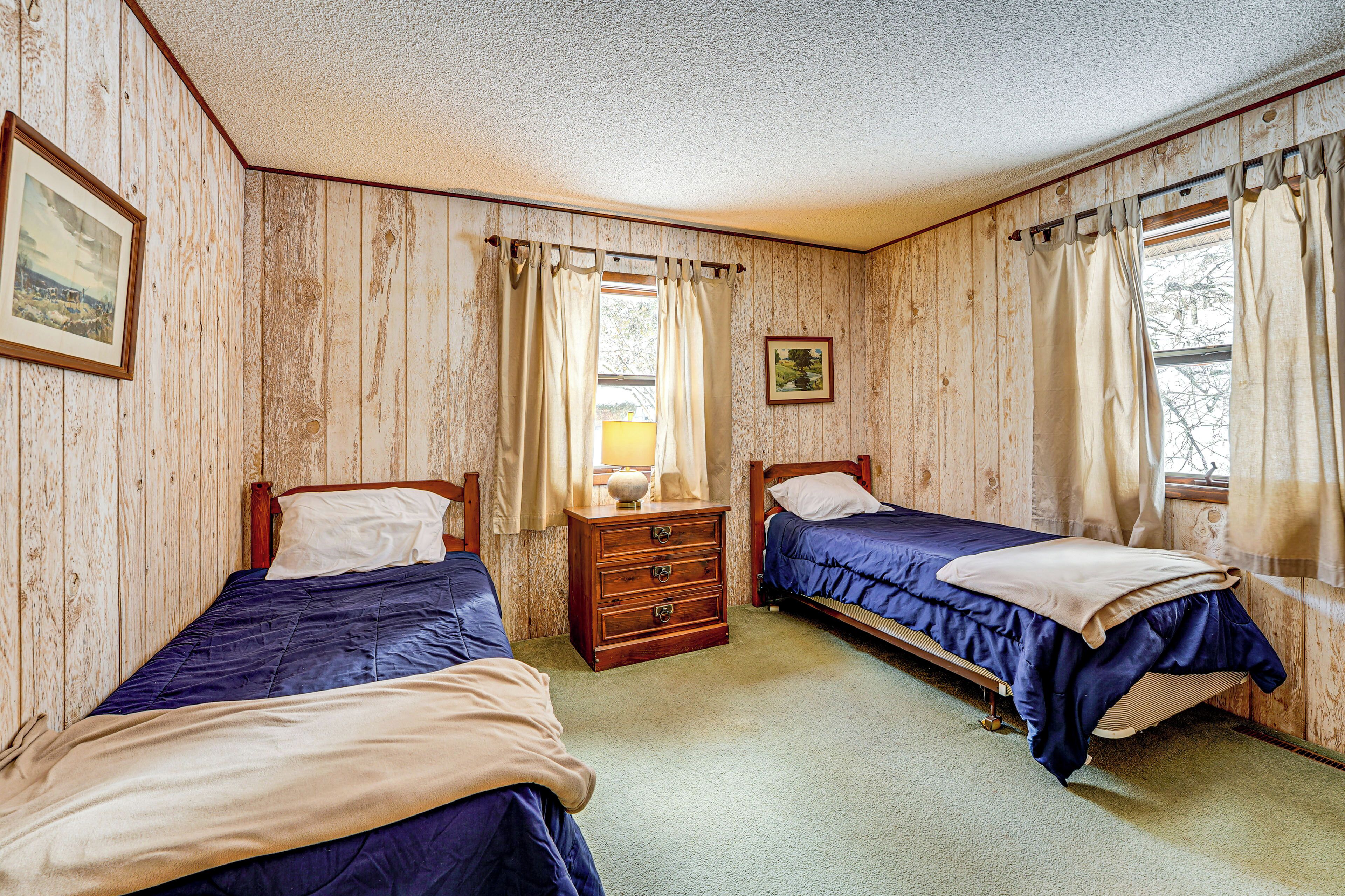 A room with two beds, wooden walls, a dresser, and a window with curtains.
