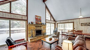 Un salon chaleureux avec une cheminée, des meubles en bois et de grandes fenêtres donnant sur l’extérieur.