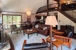 A spacious living room with a large sofa, a coffee table, and a dining area with chairs. A staircase leads to an upper level. - Pere Marquette Riverfront Cabin w/ Fishing Access (Branch)