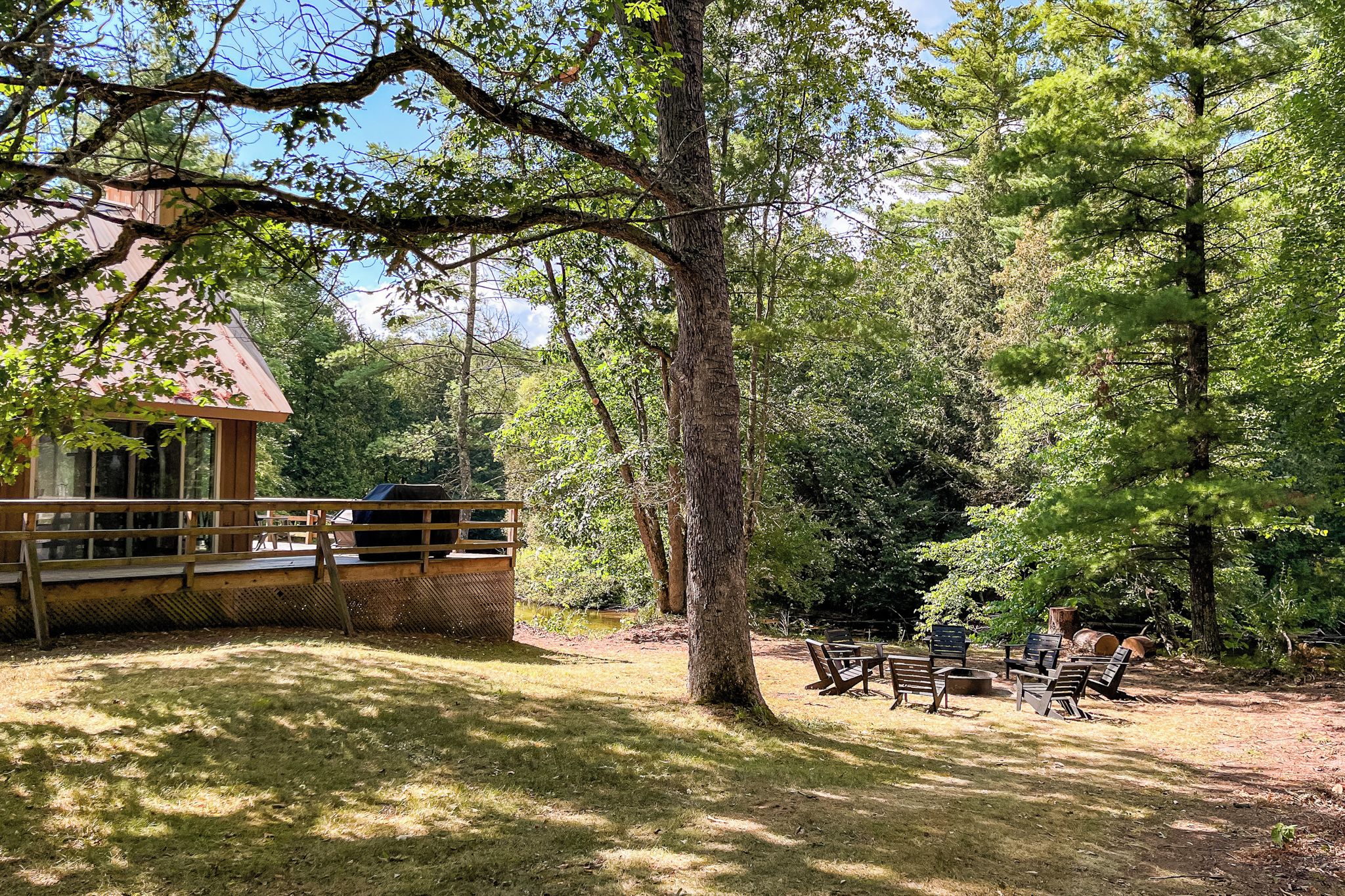 A wooden cabin with a deck, surrounded by trees and a grassy area with benches.
