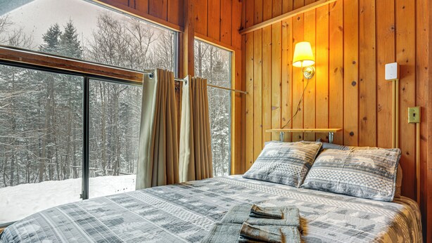 A bedroom with a large window, wooden walls, a bed with a patterned bedspread, two pillows, and a bedside lamp. - Outdoor Adventure Hub! Family Home in West Dover (West Dover)