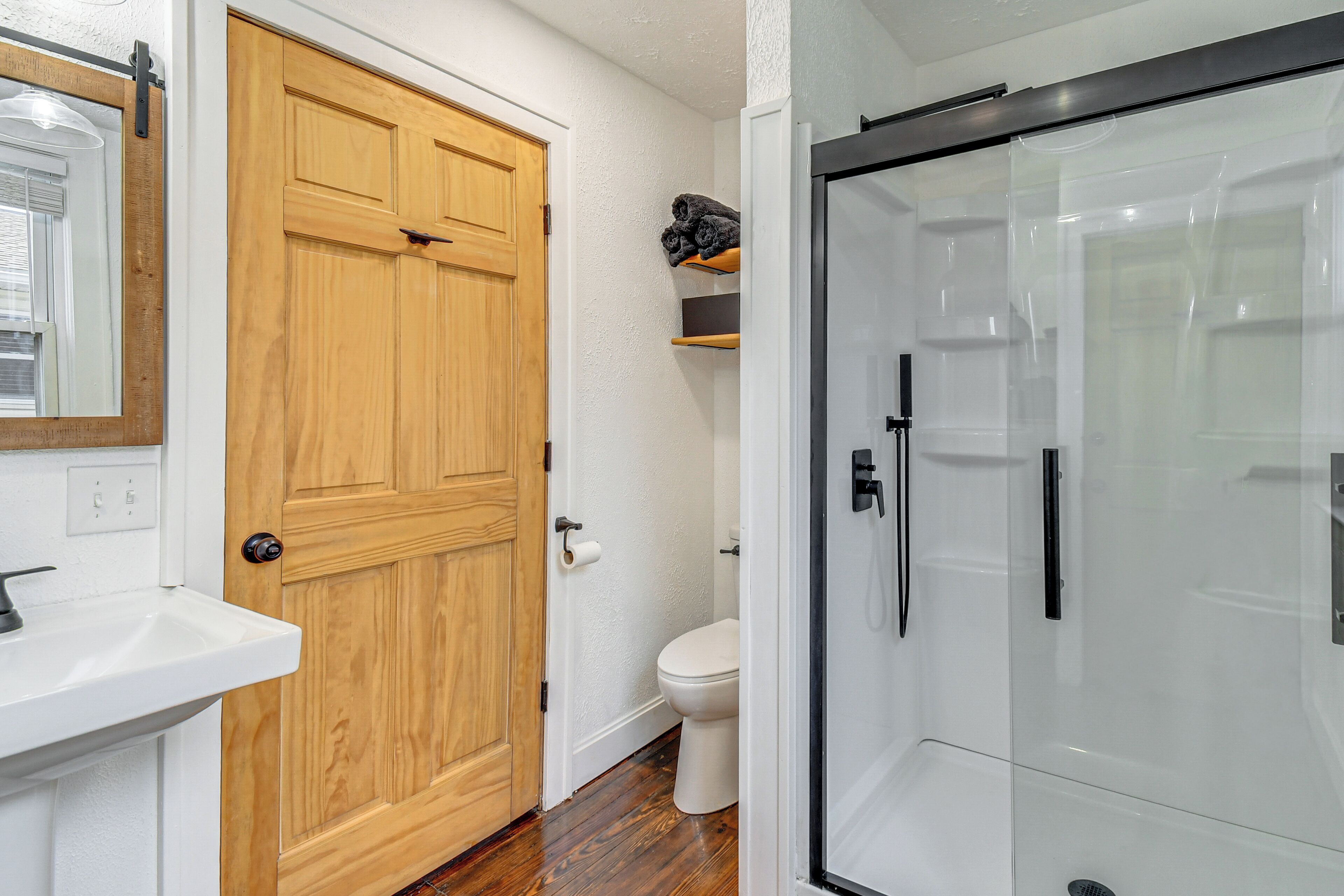 A bathroom with a wooden door, a white sink, a toilet, and a glass shower enclosure.