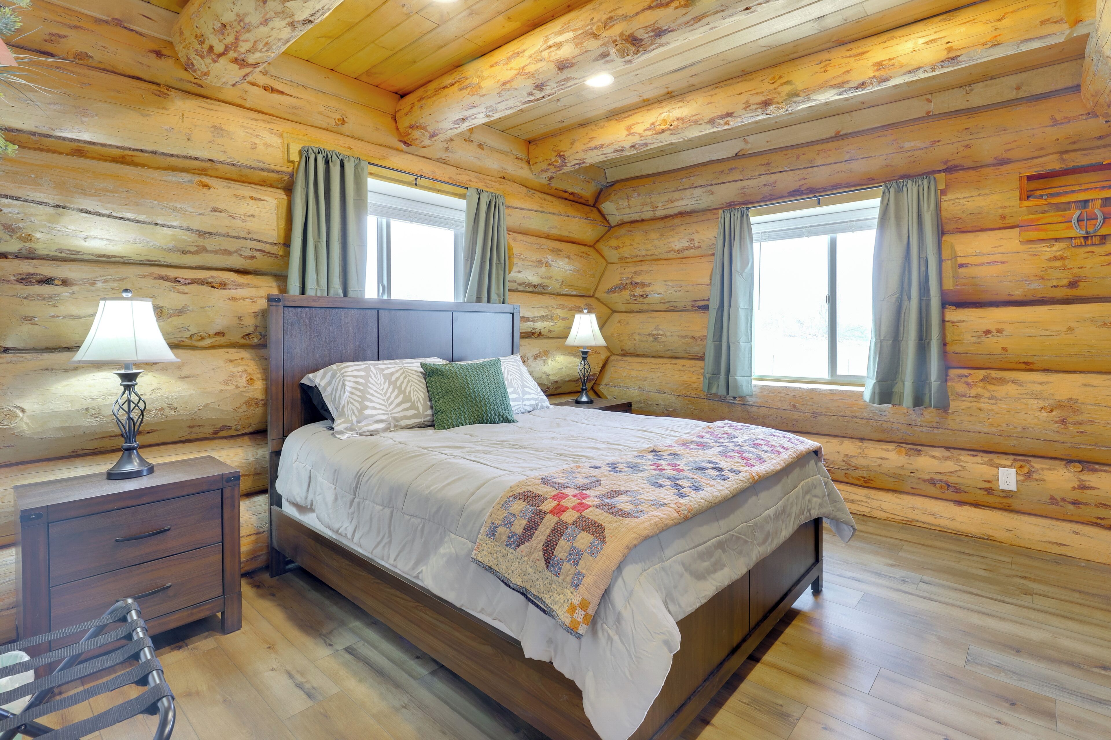 A log cabin bedroom with a wooden bed, two bedside tables with lamps, a wooden nightstand, and a window with curtains.