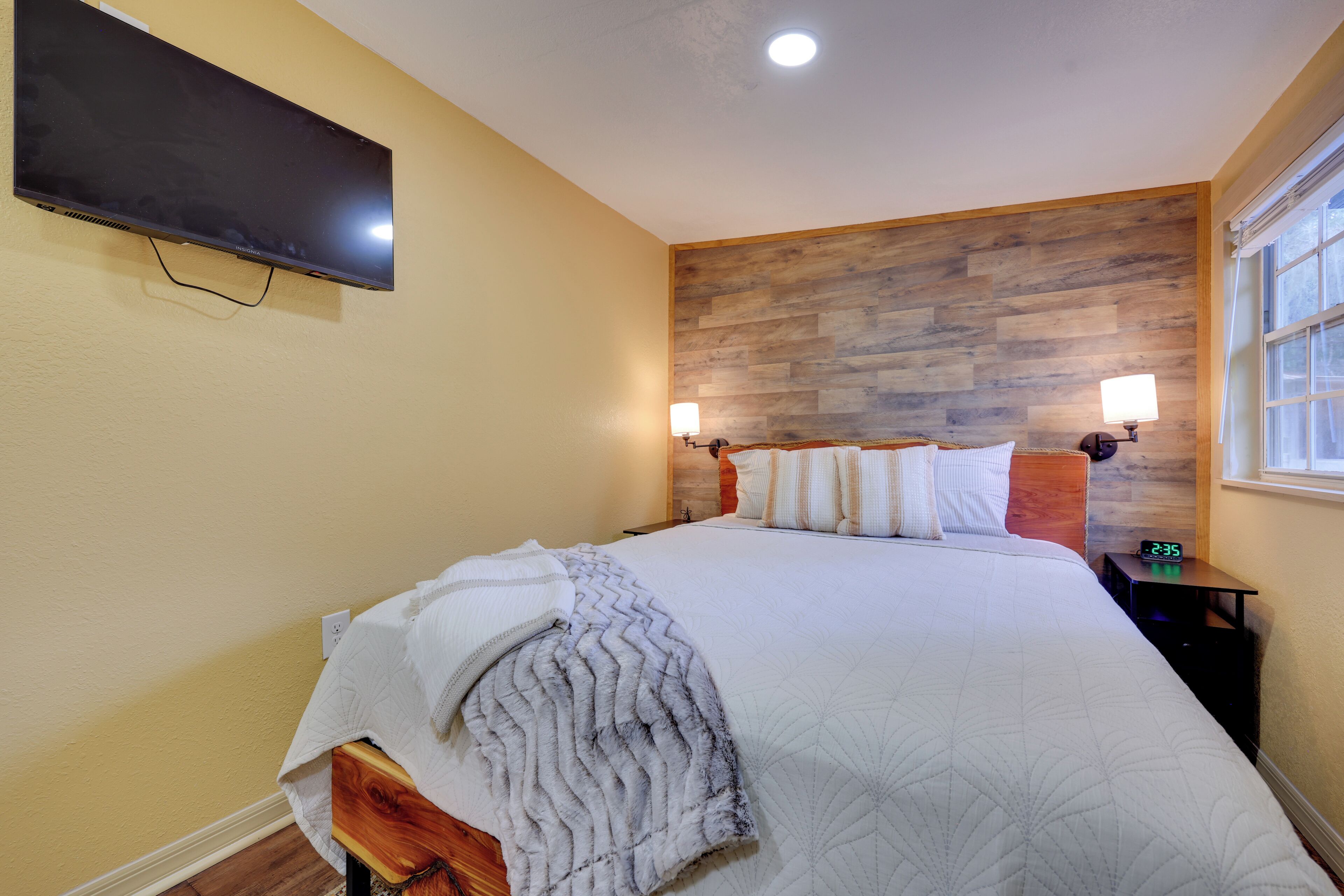 A bedroom with a large bed, a mounted TV, a wooden headboard, bedside lamps, and a window with a view.