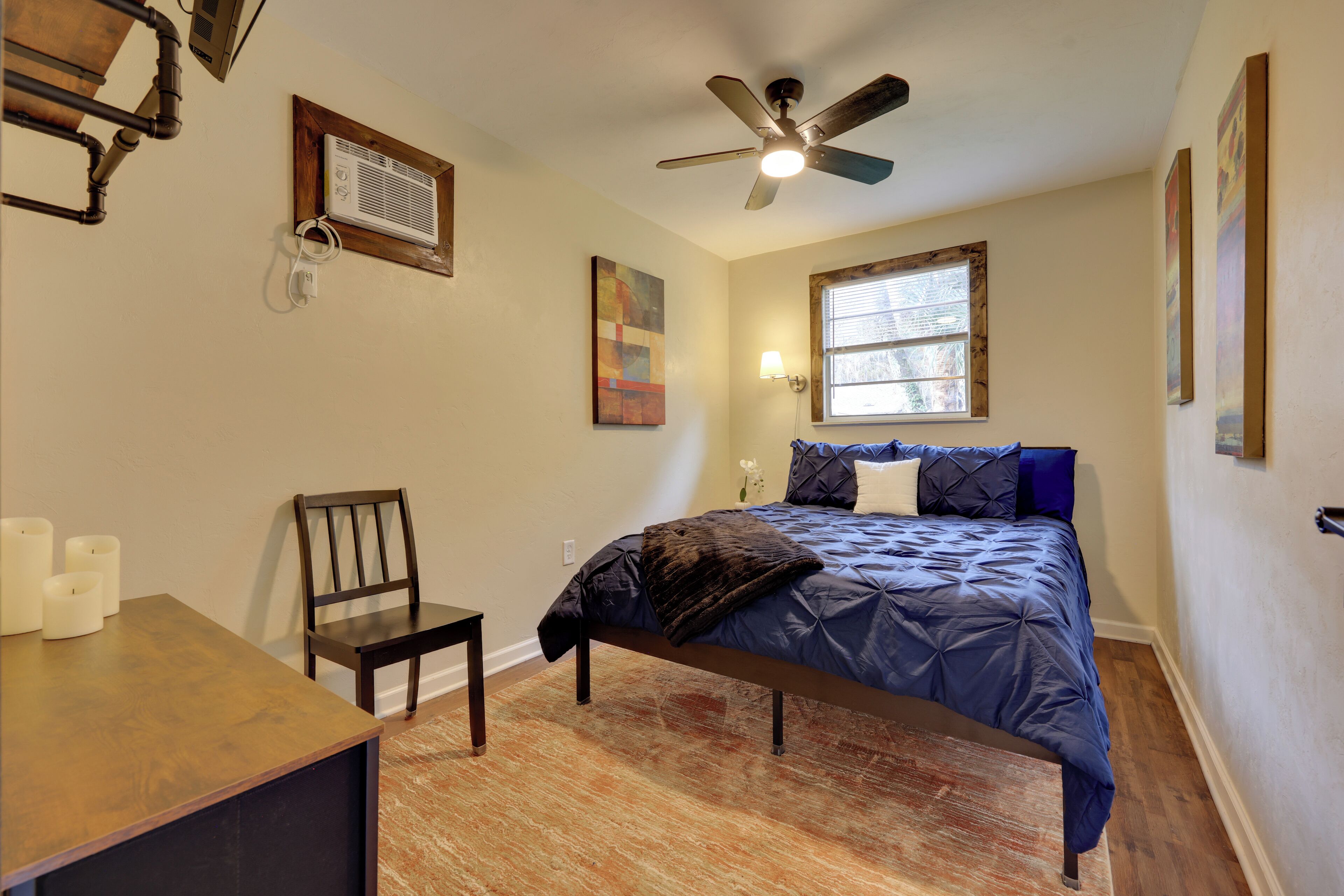 Une chambre à coucher comprenant un lit, une chaise, un bureau, un ventilateur de plafond, une fenêtre et un climatiseur fixé au mur.