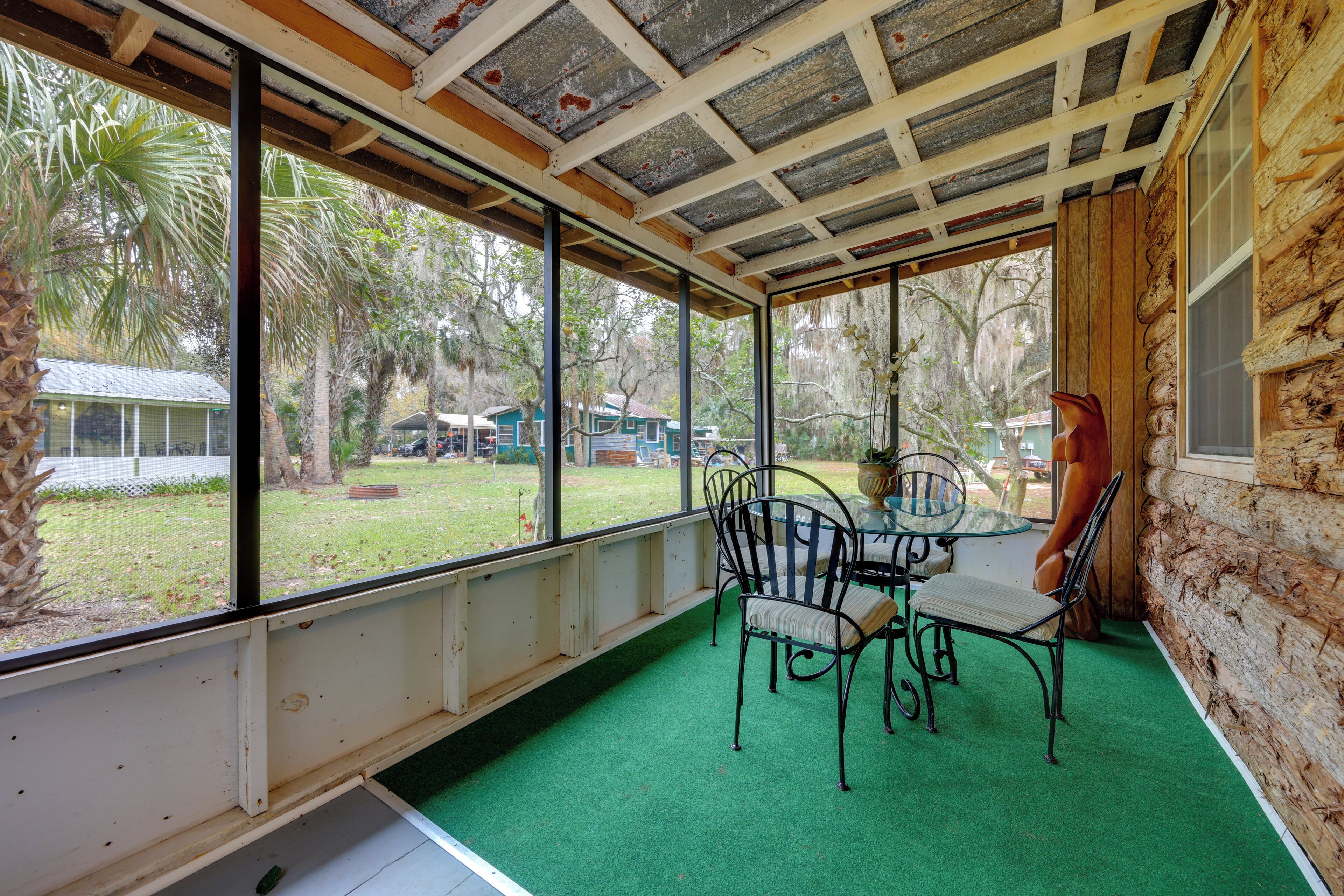 A covered patio with a table and chairs, large windows, and a view of a yard with trees and a building.