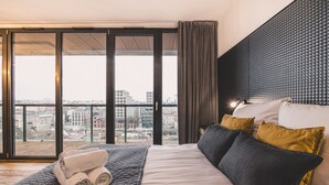 Une chambre d’hôtel avec un grand lit, une vue sur la ville grâce à de grandes fenêtres du sol au plafond et un balcon.