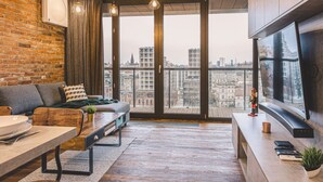 A modern living room with a sofa, a dining table, and a large window offering a city view. - Ladrillo - Luksusowy Loft z Widokiem na Miasto (Bydgoszcz)