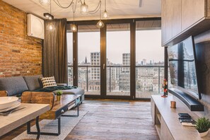 A modern living room with a sofa, a dining table, and a large window offering a city view. - Ladrillo - Luksusowy Loft z Widokiem na Miasto (Bydgoszcz)