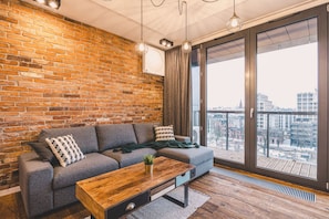 A modern living room with a brick wall, a grey sofa, a wooden coffee table, and a large window with a city view. - Ladrillo - Luksusowy Loft z Widokiem na Miasto (Bydgoszcz)