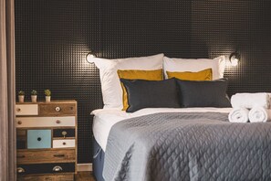 A neatly made bed with a quilted cover, a wooden nightstand with drawers, and a wall with a textured pattern. - Ladrillo - Luksusowy Loft z Widokiem na Miasto (Bydgoszcz)