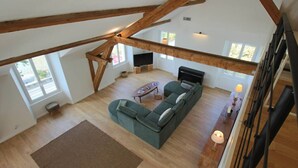 A spacious loft with high ceilings, exposed wooden beams, a large sectional sofa, a flat-screen TV, and a staircase.