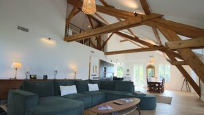 A spacious living area with a high ceiling and exposed wooden beams, a large green sofa, a wooden coffee table, and a dining area in the background.