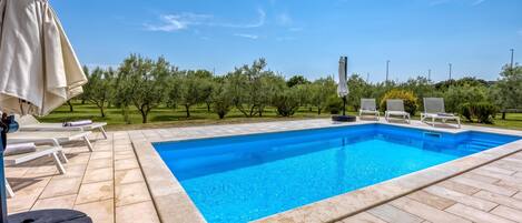 Una piscina rectangular con solárium alicatado, rodeada de sillas de descanso y sombrillas, con árboles y un cielo despejado de fondo.