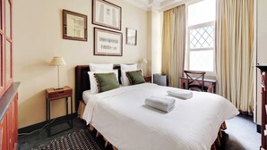 A hotel room with a large bed, two bedside tables, a desk, and a chair. There are framed pictures on the wall and a window with curtains. - Historic Elegance in the Heart of Marais - 6p/2br (Paris)
