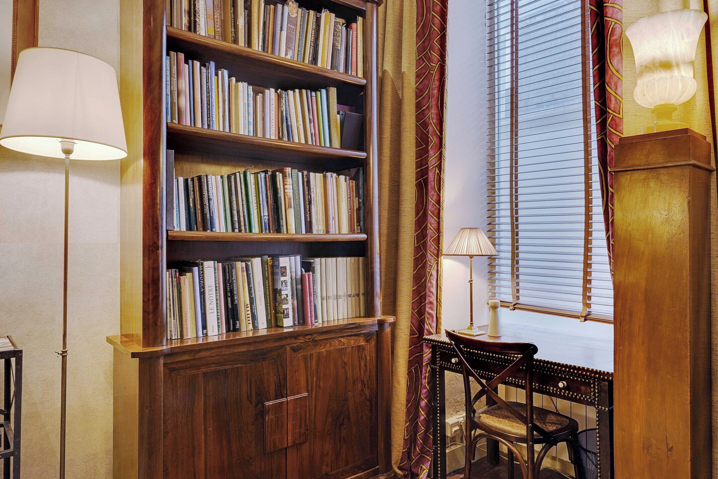 Ein Zimmer mit einem großen Bücherregal voll mit Büchern, einem Schreibtisch mit Stuhl und einem Fenster mit Jalousien.