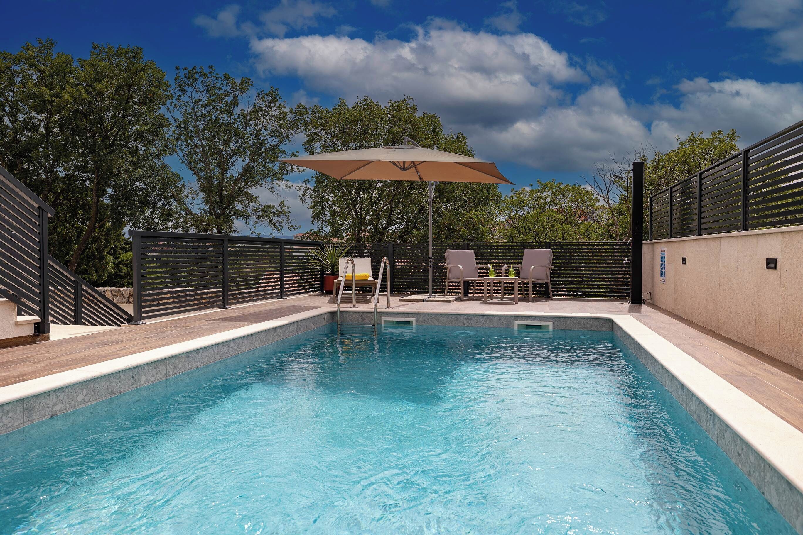 A swimming pool with a wooden deck, two lounge chairs, and a large umbrella.