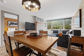 Ein zeitgemäßer Wohnraum mit Essbereich, Fernseher und großem Fenster mit Blick auf eine bewaldete Landschaft.