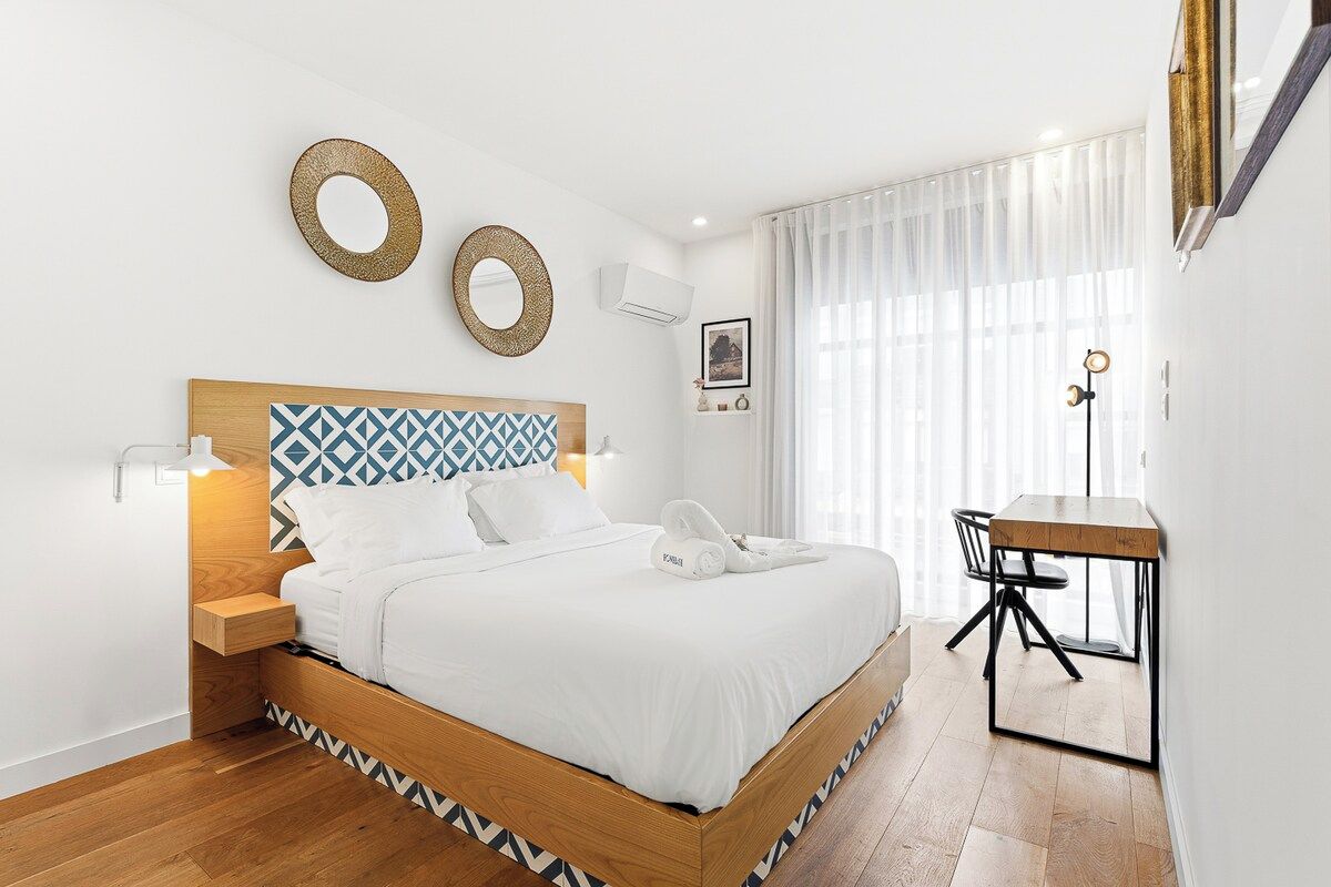 A modern bedroom with a wooden bed, bedside tables, a desk, and a large window with curtains.