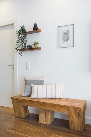 En træbænk med pude, en vægmonteret hylde med planter, og et indrammet billede.