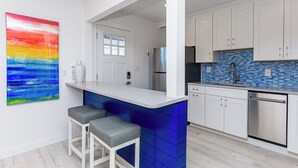 Una cocina moderna con armarios blancos, un salpicadero azul y electrodomésticos de acero inoxidable. Hay dos taburetes de bar y un cuadro abstracto y colorido en la pared.