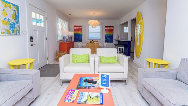 Una sala moderna con un conjunto de sofá, una mesa de centro con revistas, una zona de comedor con mesa y sillas, y una tabla de surf en la pared.